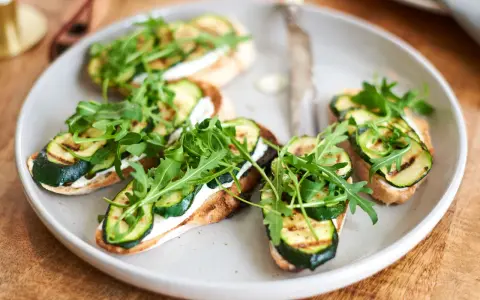 Italienische Crostini mit gegrillter Zucchini, Frischkäse und Rucola auf einem Teller.