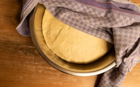 Frischer pizzateig für 4 Personen ruht in einer Schüssel, abgedeckt mit einem karierten Tuch.