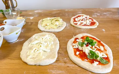 Vier vorbereitete Pinsa-Teiglinge liegen auf einem Holztisch. Einer mit Apfelscheiben, einer mit Tomatensoße und Mozzarella, einer nur mit Mozzarella und einer mit Tomatensoße, Mozzarella und Basilikum.