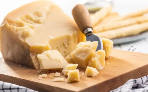 Ein Stück Parmesan mit grob abgebrochenen Stücken und einem Käsemesser. Die Parmesan Rinde ist sichtbar.