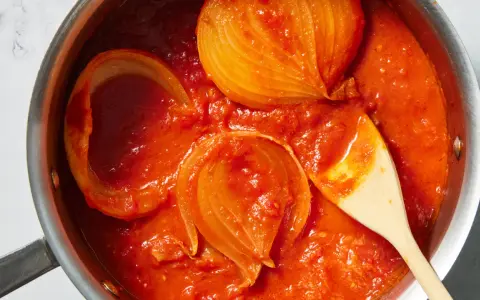 Tomatensoße mit Zwiebelstücken in einem Topf, bereit für Marcella Hazan Rezepte. Ein Holzlöffel rührt die köstliche Soße.