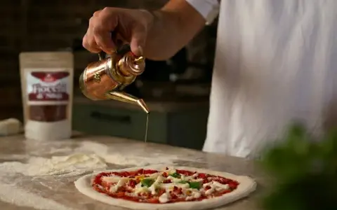 Ein Mann übergießt mit Olivenöl einen **Waldi Pizzateig**. Im Hintergrund eine Tüte mit Chiliflocken.