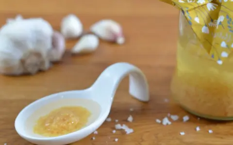 Ein Löffel mit einer köstlichen Knoblauchpaste, die an italienische Aromen erinnert. Daneben eine Flasche mit einer gelben Flüssigkeit und Knoblauchzehen.