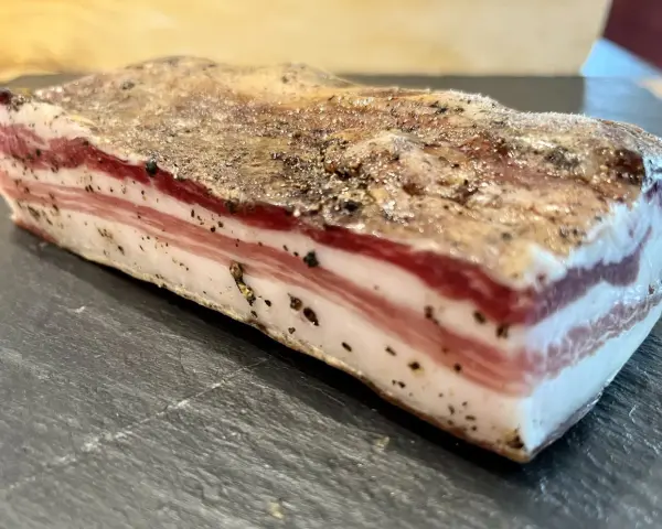 Ein Stück roher Pancetta mit deutlichen Schichten aus Fleisch und Fett, bestreut mit schwarzem Pfeffer. Perfekt, um ihn roh zu genießen.