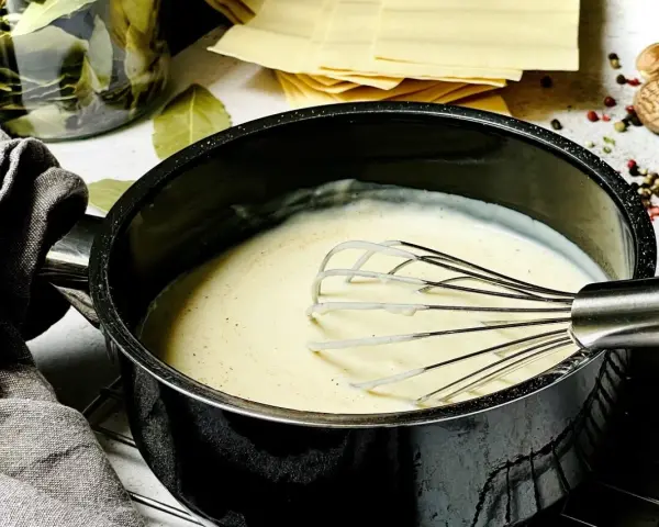 Cremige béchamelsauce mit Sahne wird in einem Topf mit einem Schneebesen zubereitet. Daneben liegen Lasagneplatten und Gewürze.
