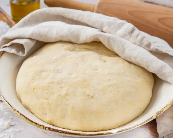 Der weltbeste Pizzateig mit Trockenhefe ruht abgedeckt in einer Schüssel. Daneben liegen Salz, Olivenöl und ein Nudelholz.