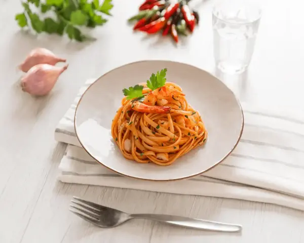 Ein Teller mit Linguini Nudeln mit Garnelen und Tomatensauce, garniert mit Petersilie. Daneben Schalotten, Chilis und ein Glas Wasser.