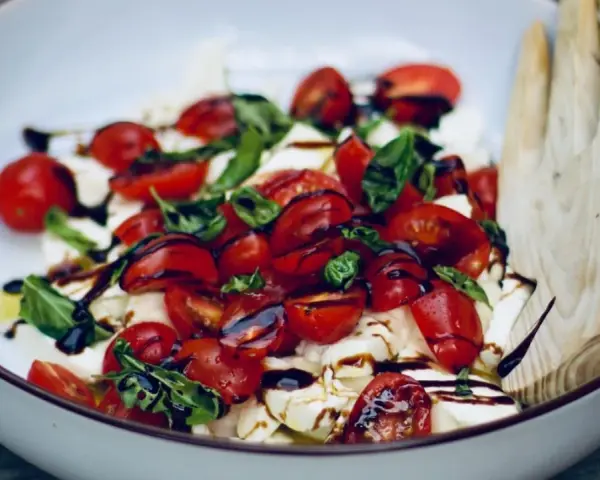 Burrata Caprese Salat mit Kirschtomaten, Basilikum und Balsamico-Dressing, serviert in einer Schüssel mit Holzsalatbesteck.
