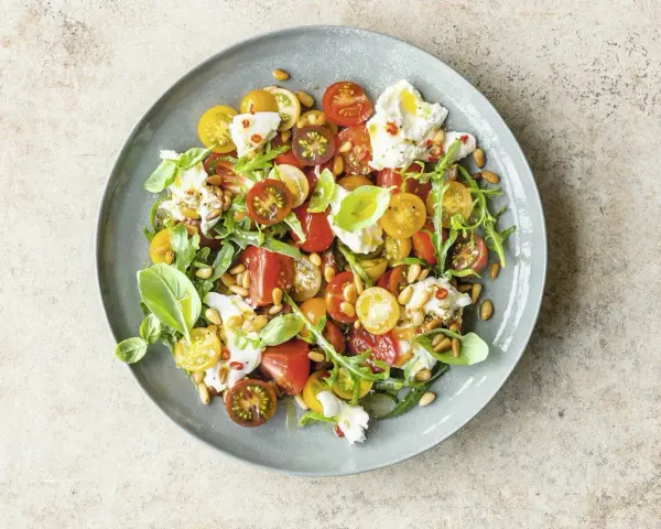 Bunter Tomatensalat mit cremiger Burrata, Rucola und Pinienkernen, garniert mit Basilikum.