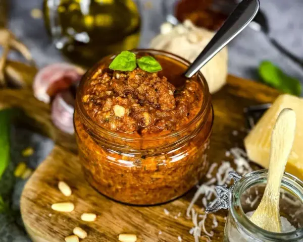 Ein Glas mit leuchtend rotem Pesto, garniert mit Basilikum. Daneben liegen Pinienkerne, Knoblauch und ein Stück Parmesan.