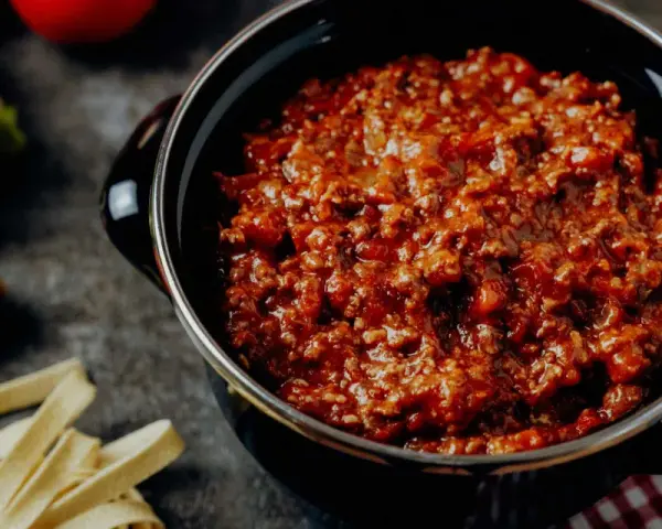 Herzhafte Bolognese-Sauce in einem Topf, daneben frische Bolognese-Zutaten wie Karotten, Sellerie und Tomaten.