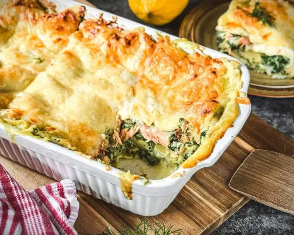Eine köstliche, goldbraun überbackene italienische Lachslasagne mit Spinat und cremiger Sauce. Ein Stück ist bereits serviert.