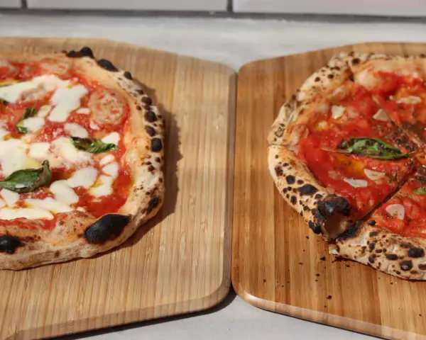 Zwei frisch gebackene Pizzen auf Holzbrettern, eine Margherita, die andere mit Tomaten und Basilikum.
