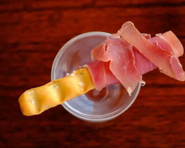 Ein knuspriger Grissini, umwickelt mit zartem Schinken, ruht in einem Glas. Perfekt für den kleinen Hunger.