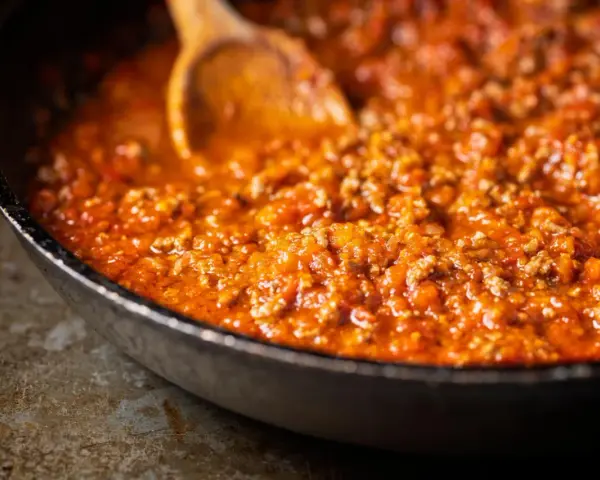 Ein Holzlöffel rührt in einer Pfanne mit köchelndem Sos bolognese.
