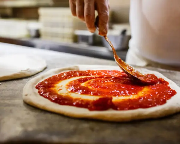Eine Hand verteilt Tomatensauce auf einem Teigboden, die Vorbereitung für eine authentische pizza napoli original.