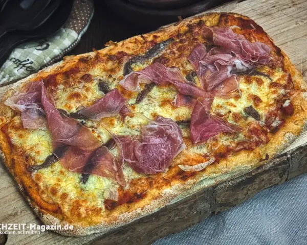Eine rechteckige Pinsa mit geschmolzenem Käse und hauchdünnen Scheiben Schinken belegen. Köstlich!