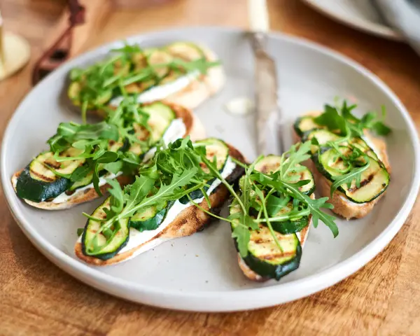 Italienische Crostini mit gegrillter Zucchini, Frischkäse und Rucola auf einem Teller.