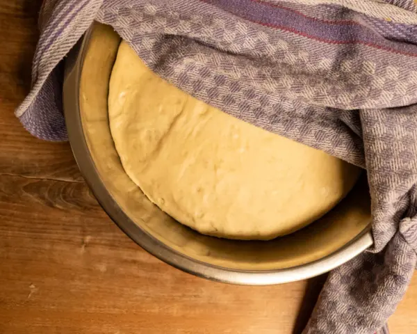 Frischer pizzateig für 4 Personen ruht in einer Schüssel, abgedeckt mit einem karierten Tuch.
