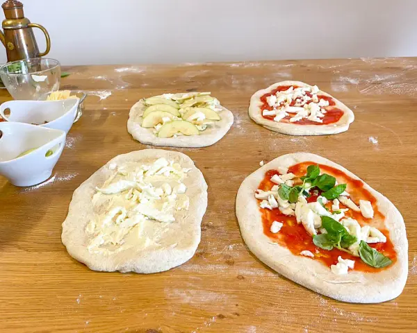 Vier vorbereitete Pinsa-Teiglinge liegen auf einem Holztisch. Einer mit Apfelscheiben, einer mit Tomatensoße und Mozzarella, einer nur mit Mozzarella und einer mit Tomatensoße, Mozzarella und Basilikum.