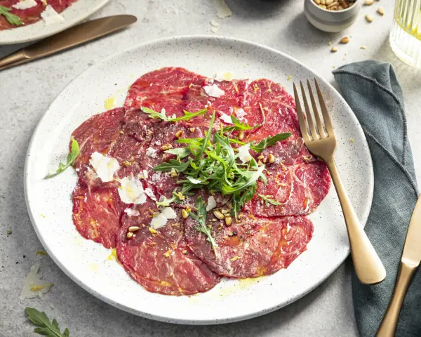 Carpaccio selber machen: hauchdünne Rinderfiletscheiben mit Rucola, Parmesan und Pinienkernen auf einem Teller.