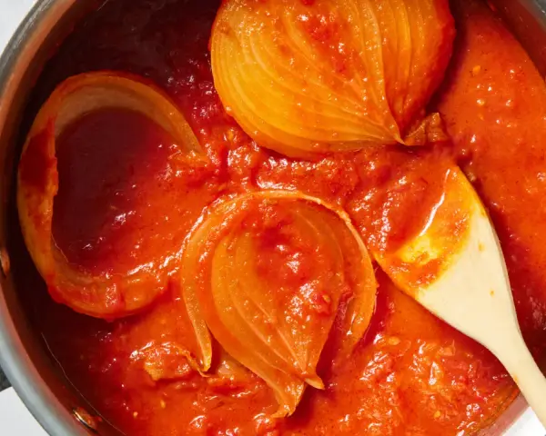 Tomatensoße mit Zwiebelstücken in einem Topf, bereit für Marcella Hazan Rezepte. Ein Holzlöffel rührt die köstliche Soße.