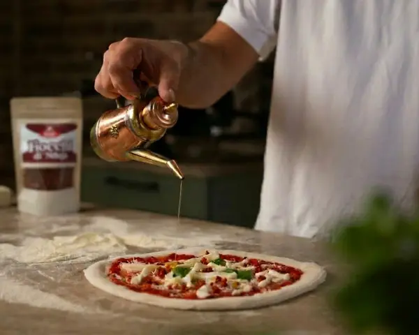 Ein Mann übergießt mit Olivenöl einen **Waldi Pizzateig**. Im Hintergrund eine Tüte mit Chiliflocken.