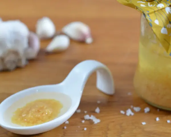 Ein Löffel mit einer köstlichen Knoblauchpaste, die an italienische Aromen erinnert. Daneben eine Flasche mit einer gelben Flüssigkeit und Knoblauchzehen.