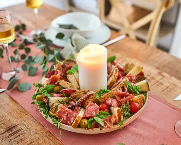 Ein festliches Arrangement mit italienischen Snacks zum Wein: Salami, Prosciutto, Käse, Oliven und Brot, umrahmt von Kräutern und einer Kerze.