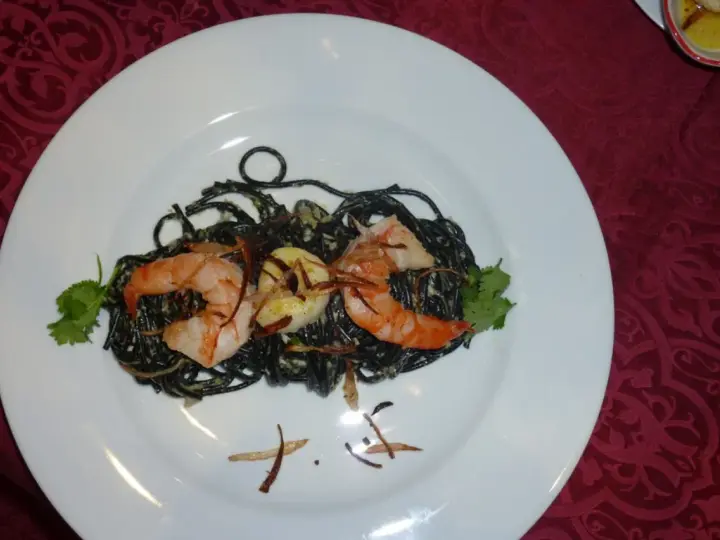 Schwarze Spaghetti mit Garnelen, garniert mit Koriander und knusprigen Zwiebeln. Ein köstliches Gericht, das auf einem weißen Teller serviert wird.