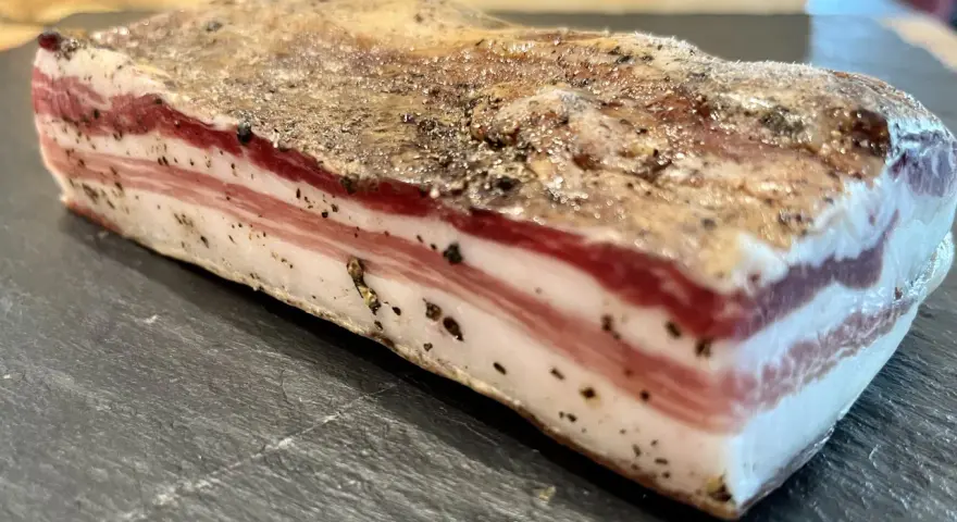Ein Stück roher Pancetta mit deutlichen Schichten aus Fleisch und Fett, bestreut mit schwarzem Pfeffer. Perfekt, um ihn roh zu genießen.