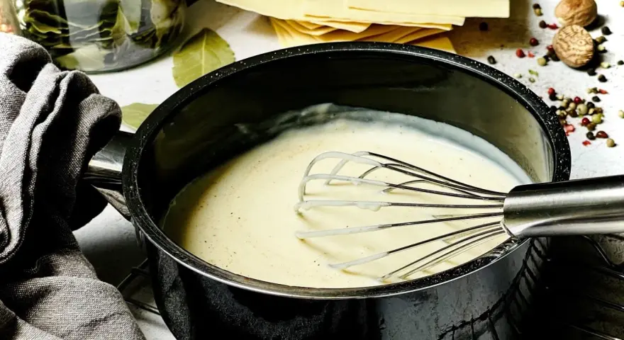 Cremige béchamelsauce mit Sahne wird in einem Topf mit einem Schneebesen zubereitet. Daneben liegen Lasagneplatten und Gewürze.