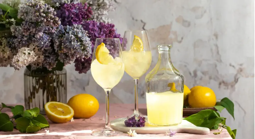 Erfrischender Limoncello Spritz mit Bitter Lemon, garniert mit Zitronenspalten, neben frischen Zitronen und einem üppigen Fliederstrauß.