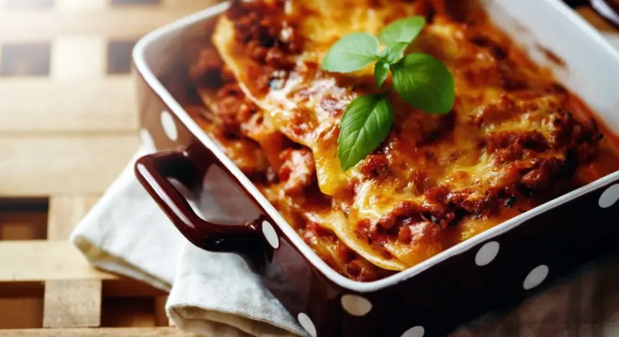 Herzhafte Lasagne Bolognese, eine der beliebtesten Lasagne Arten, dampft in einer braunen Auflaufform mit weißen Punkten. Frisch garniert mit Basilikum.