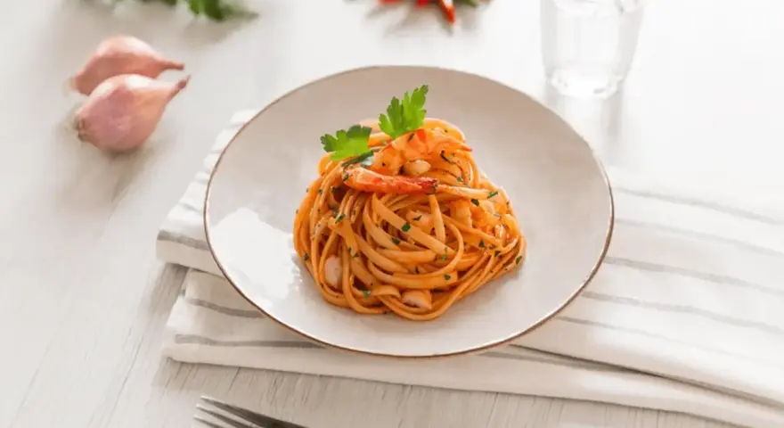 Ein Teller mit Linguini Nudeln mit Garnelen und Tomatensauce, garniert mit Petersilie. Daneben Schalotten, Chilis und ein Glas Wasser.