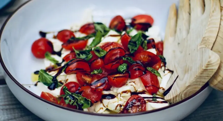 Burrata Caprese Salat mit Kirschtomaten, Basilikum und Balsamico-Dressing, serviert in einer Schüssel mit Holzsalatbesteck.