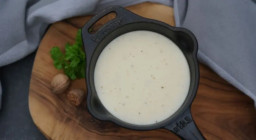 Cremige Bechamelsauce in einer Gusseisenpfanne, bereit für die Lasagne. Muskatnuss und Petersilie deuten auf die feinen Aromen hin.