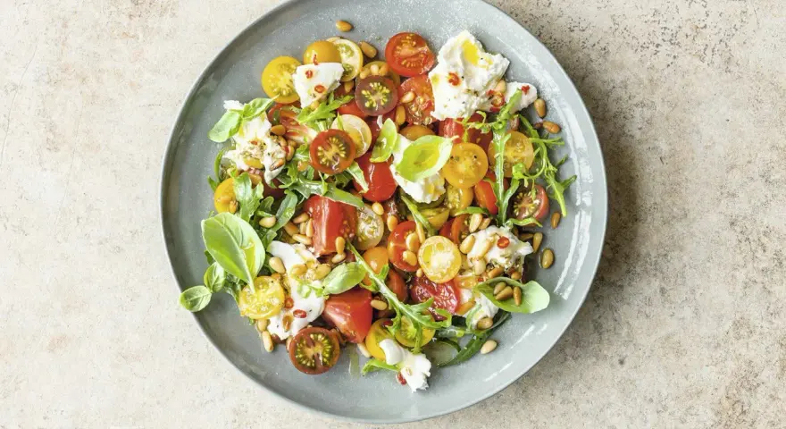 Bunter Tomatensalat mit cremiger Burrata, Rucola und Pinienkernen, garniert mit Basilikum.