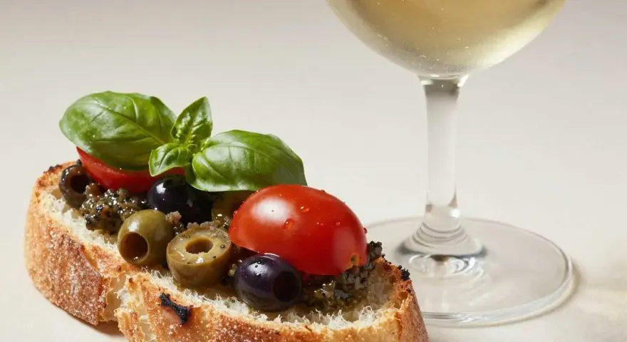 Bruschetta mit Olivencreme, Tomaten und Basilikum. Ein Glas Weißwein daneben. Perfekt für ein schnelles olivencreme rezept.