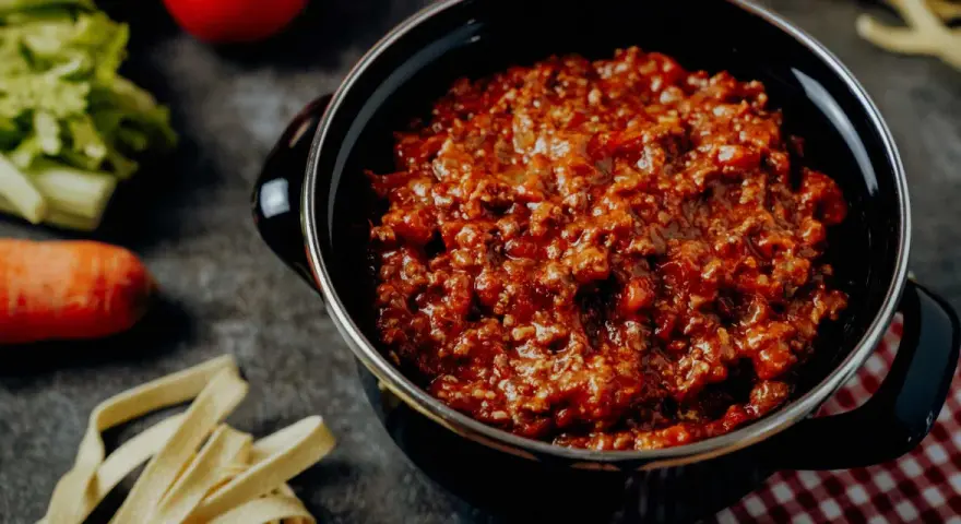 Herzhafte Bolognese-Sauce in einem Topf, daneben frische Bolognese-Zutaten wie Karotten, Sellerie und Tomaten.