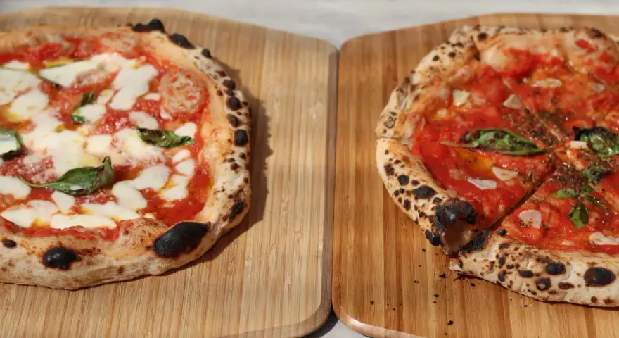 Zwei frisch gebackene Pizzen auf Holzbrettern, eine Margherita, die andere mit Tomaten und Basilikum.