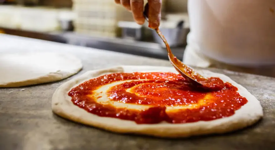 Eine Hand verteilt Tomatensauce auf einem Teigboden, die Vorbereitung für eine authentische pizza napoli original.
