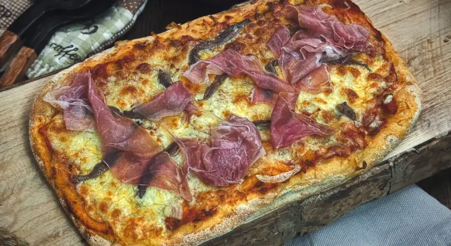 Eine rechteckige Pinsa mit geschmolzenem Käse und hauchdünnen Scheiben Schinken belegen. Köstlich!