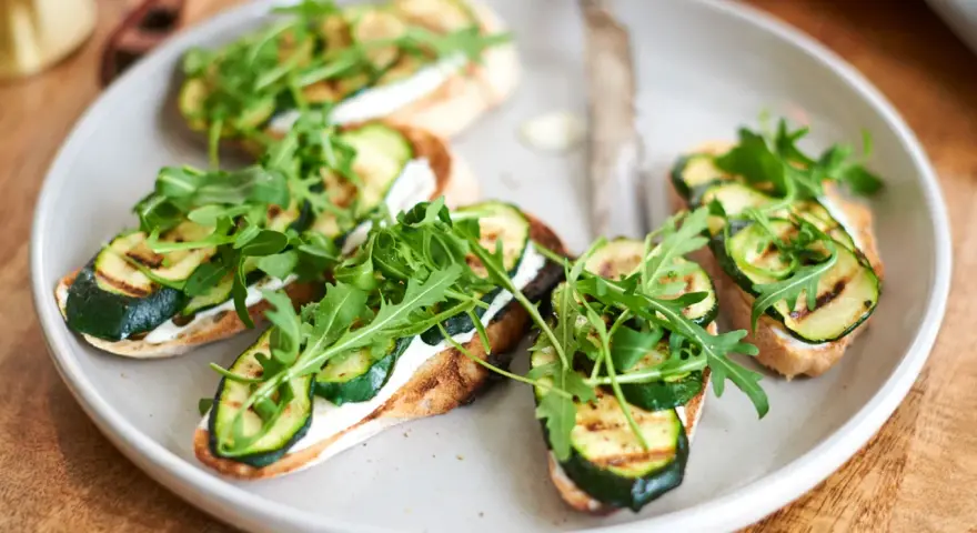 Italienische Crostini mit gegrillter Zucchini, Frischkäse und Rucola auf einem Teller.