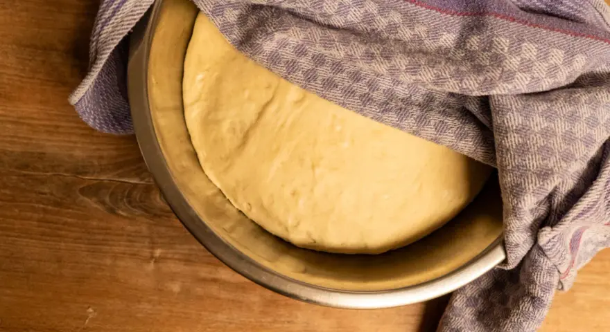 Frischer pizzateig für 4 Personen ruht in einer Schüssel, abgedeckt mit einem karierten Tuch.