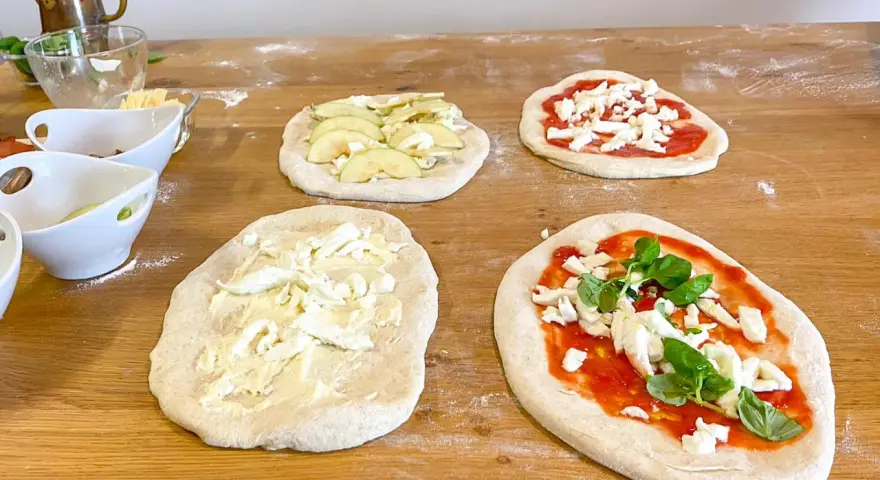 Vier vorbereitete Pinsa-Teiglinge liegen auf einem Holztisch. Einer mit Apfelscheiben, einer mit Tomatensoße und Mozzarella, einer nur mit Mozzarella und einer mit Tomatensoße, Mozzarella und Basilikum.