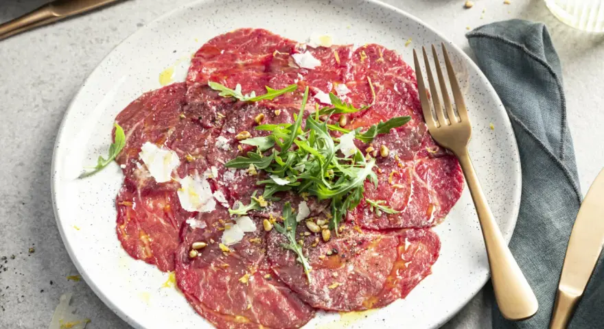 Carpaccio selber machen: hauchdünne Rinderfiletscheiben mit Rucola, Parmesan und Pinienkernen auf einem Teller.
