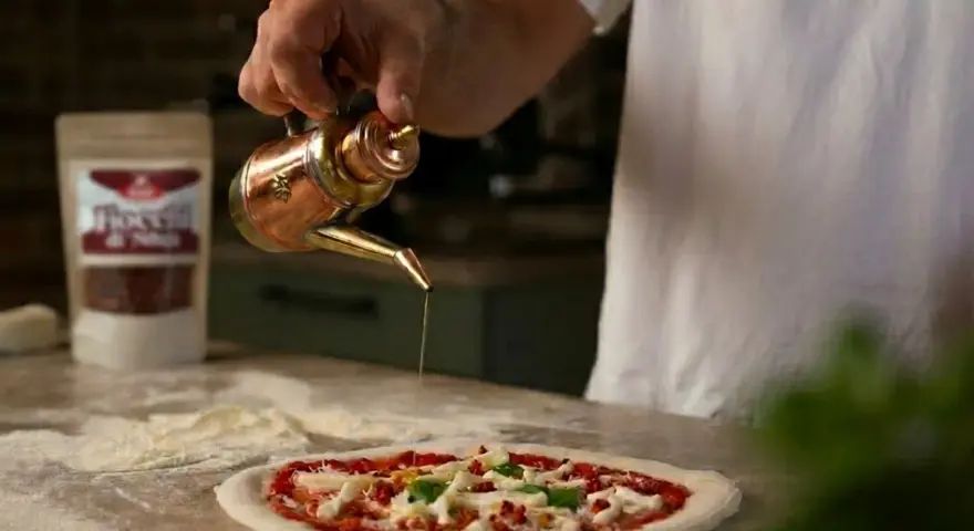 Ein Mann übergießt mit Olivenöl einen **Waldi Pizzateig**. Im Hintergrund eine Tüte mit Chiliflocken.