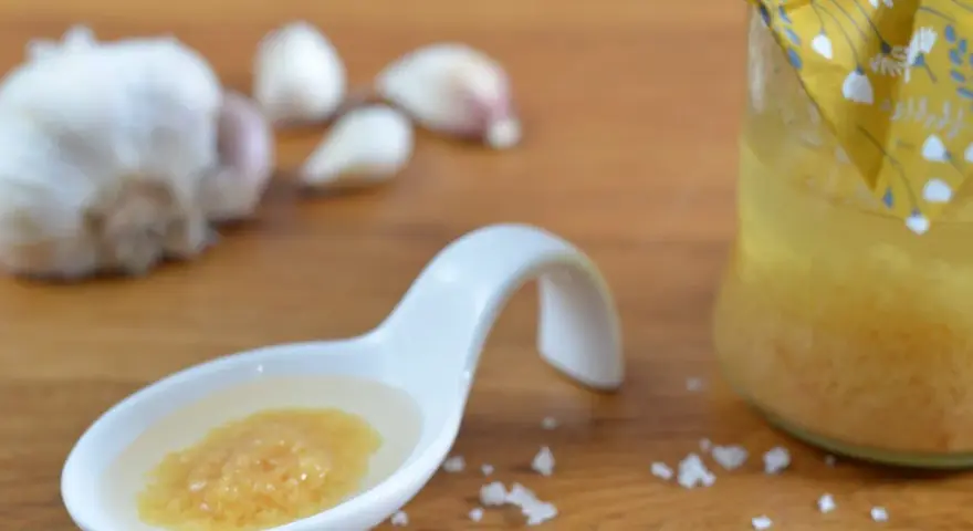 Ein Löffel mit einer köstlichen Knoblauchpaste, die an italienische Aromen erinnert. Daneben eine Flasche mit einer gelben Flüssigkeit und Knoblauchzehen.