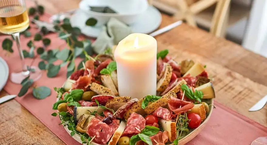 Ein festliches Arrangement mit italienischen Snacks zum Wein: Salami, Prosciutto, Käse, Oliven und Brot, umrahmt von Kräutern und einer Kerze.